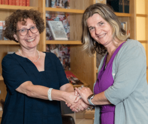 Nieuw directeur Wilma van Wezenbeek en huidig directeur Lilly Knibbeler schudden elkaar de hand, met een grote glimlach. Ze staan voor een van de boekenkasten van de KB.